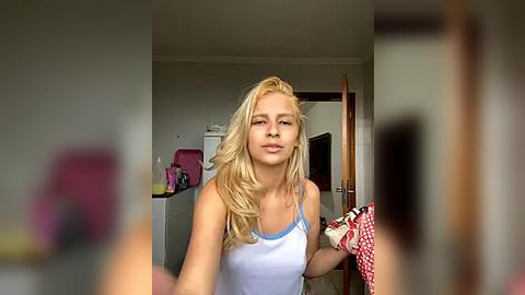 Media: Video of a blonde woman in a light blue tank top, standing indoors in a cluttered room with a red checkered cloth and a pink bag in the background.