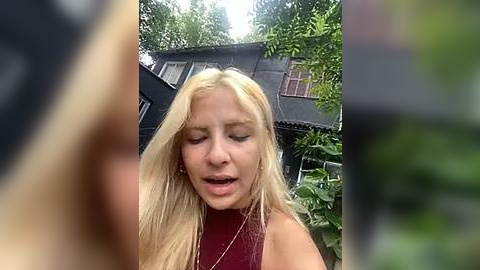 Media: A candid video of a blonde woman with long hair, wearing a sleeveless top, outside near a dark wooden house with green foliage. She appears to be in distress, with her hand on her head, mouth open.