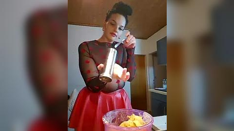 Media: Video of a woman with dark hair in a bun, wearing a sheer black top with red polka dots and a red skirt, stirring a bowl of yellow batter.