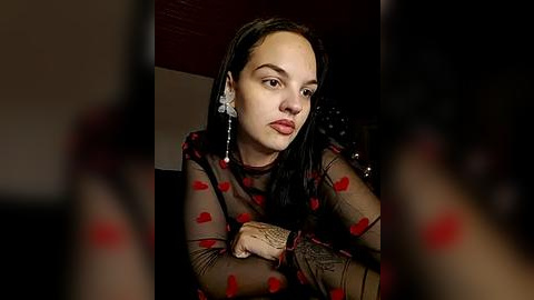 Media: Video of a woman with medium skin tone and long black hair, wearing a sheer black dress with red heart patterns and silver earrings, looking contemplative.