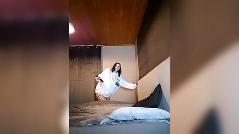 Media: A video captures a young woman in a white hoodie, standing in a dimly lit, minimalist bedroom with beige walls, wooden ceiling, and a bed with white sheets.