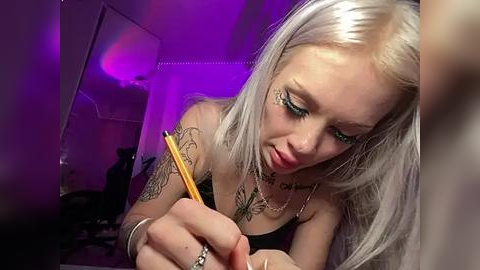 Media: Video of a young, platinum blonde woman with blue eyes and intricate tattoos, writing with a pencil in a dimly lit room under purple lighting.