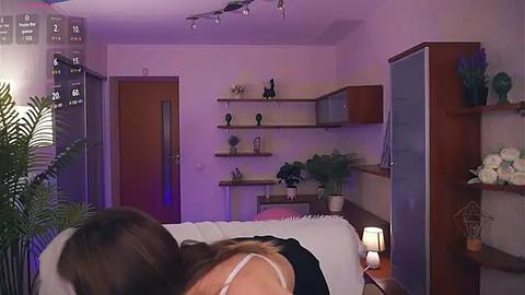 Media: A video of a cozy, dimly lit bedroom with a woman lying face down on a bed, wearing a black dress and a white blanket. The room features wooden furniture, potted plants, and a purple light.