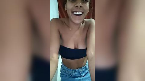 Media: Video of a young woman with light brown skin, wearing a black strapless top and high-waisted denim shorts, grinning widely.