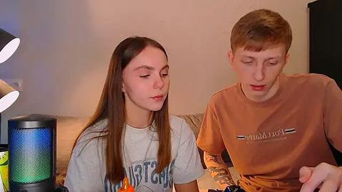 Media: Video of a young woman with long brown hair and a young man with short red hair, both in casual T-shirts, playing video games on a couch with a colorful lamp and a TV in the background.