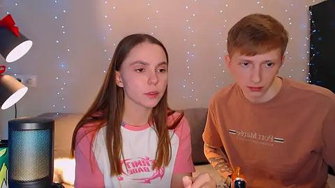 Media: Video of a young Caucasian girl with long brown hair, wearing a pink and white T-shirt, and a boy with short brown hair, both sitting indoors with a cozy, dimly lit background.