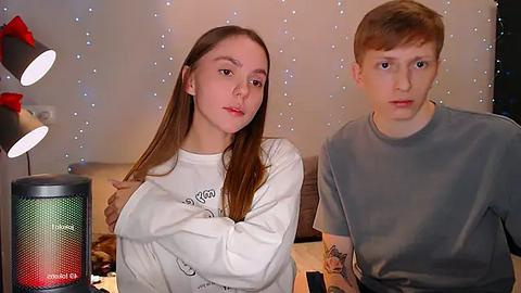 Media: Video of two young, fair-skinned individuals with straight brown hair, wearing white and grey t-shirts, seated indoors with a green and red 'Cordless' speaker and decorative lights in the background.