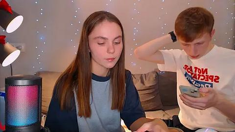 Media: Video of two young people: a brunette girl in a blue shirt, a fair-skinned boy in a white shirt with \"PAPA JONES\" logo, both engrossed in their phones, in a cozy, dimly-lit room with fairy lights and a colorful speaker.