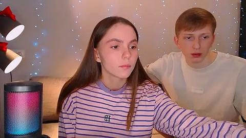 Media: Video of a young girl with long brown hair in a striped shirt, standing beside a young boy with short red hair in a white shirt. They are in a cozy room with fairy lights and a black speaker with a red bow.