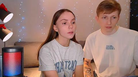 Media: Video of a young woman with long brown hair wearing a grey T-shirt with \"Patriot\" text, and a young man with short red hair in a white T-shirt, sitting on a beige couch, with a red and blue cylindrical speaker and string lights in the background.