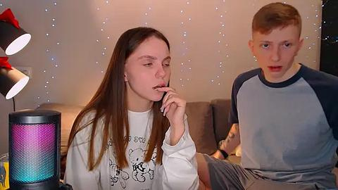 Media: Video of a young woman with long brown hair, wearing a white shirt, and a young man with short red hair in a gray shirt, both looking at a colorful Bluetooth speaker.