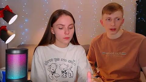 Media: Video of a teenage girl with long brown hair, wearing a white shirt with cartoon bears, and a boy with short red hair in a brown shirt. Background has string lights, a black lamp, and a blue and white can.