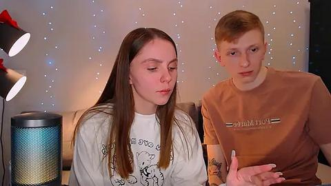 Media: Video of a young Caucasian couple with fair skin and long hair, sitting indoors. He wears a brown shirt, she a white tee. Background features a twinkling light panel and a modern lamp.