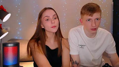 Media: Video of a young Caucasian couple with long hair, seated indoors, wearing casual black and white T-shirts, with a colorful speaker and background lights.