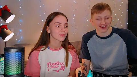 Media: Video of a young woman with long brown hair in a pink and white \"Team America\" T-shirt and a young man with short red hair in a blue and grey shirt, both smiling, in a cozy room with string lights and a speaker.