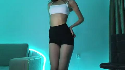 Media: Video of a slender, fair-skinned woman in a white sports bra and black shorts, standing confidently against a teal-lit background, featuring a beige sofa and a dark wooden chair.