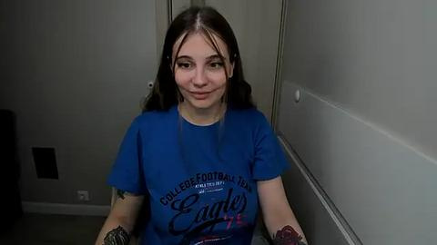 Media: Video of a young woman with fair skin and long dark hair, wearing a blue t-shirt with \"Eagle Creek\" text. She has tattoos on both arms, standing indoors in a narrow hallway with gray walls.