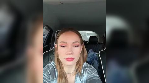 Media: Video of a young woman with light skin, straight blonde hair, and pink eyeshadow, wearing a silver velvet jacket, seated in a car with blurry passengers in the background.