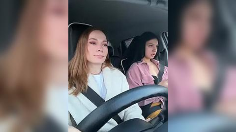 Media: Video of two women in a car, one with long, straight, light brown hair, wearing a white jacket, driving; the other with long, straight, black hair, wearing a pink shirt, looking serious.