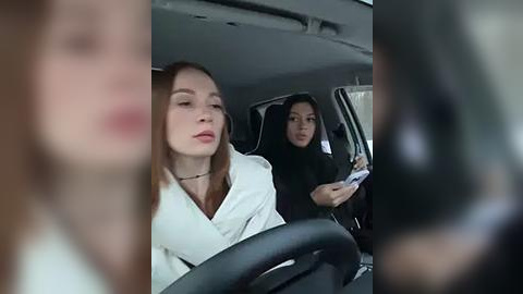 Media: Video of a woman with light skin and straight brown hair wearing a white shirt driving a car, with a woman with dark skin and long black hair in the passenger seat.