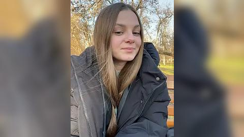 Media: Video of a young woman with long brown hair, wearing a black puffer jacket, sitting outdoors in a park with autumn foliage in the background.