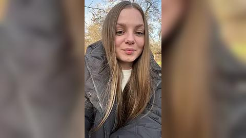 Media: A video of a young woman with straight, light brown hair and fair skin, wearing a black puffy jacket and a white shirt. The background features autumn trees and a clear blue sky, creating a serene outdoor setting.