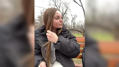 Media: Video of a young woman with long brown hair, wearing a black puffy jacket, sitting on a park bench. She holds a gray scarf with a neutral expression. Background features leafless trees and a gravel path.