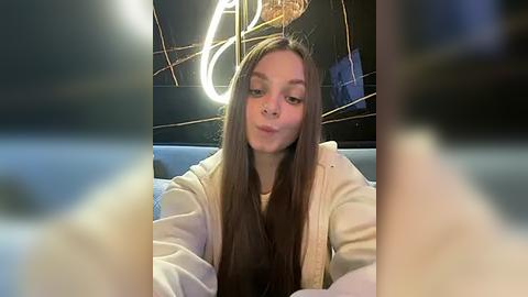 Media: Video of a young woman with long brown hair, wearing a white hoodie, sitting on a couch in a dimly lit room with a neon light.
