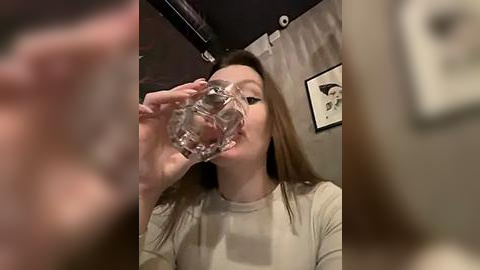 Media: A video shows a young woman with long brown hair, wearing a beige top, drinking water from a glass. The blurred background features a framed picture and a ceiling light.