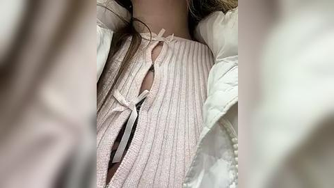 Media: Video of a woman's torso in a light pink, ribbed, high-necked blouse with a ribbon bow detail. She is wearing a white puffer jacket and has light brown hair.