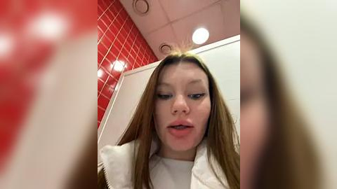 Media: A blurry video of a young woman with long hair, wearing a white jacket, standing in a red-tiled bathroom with white walls and recessed lighting.