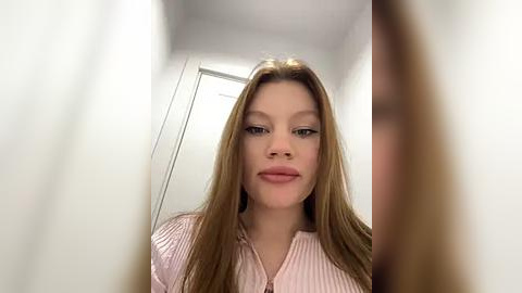 Media: Video of a young, fair-skinned woman with long, straight, light brown hair, wearing a light pink, ribbed blouse. She has a neutral expression, standing indoors with a white door and blurry background.