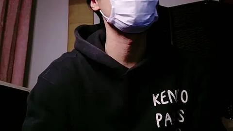 Media: Video of a person wearing a white surgical mask, black hoodie with \"Keno Paris\" printed in white, standing indoors with dim lighting.