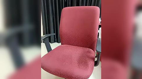 Media: Video of a red, textured office chair with a high backrest and black armrests. The background features black curtains and a blurred desk, giving a professional setting.