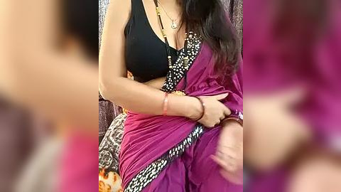 Media: Video of a woman in a black bra, purple sari with a black floral border, holding a red sari, with blurred background.