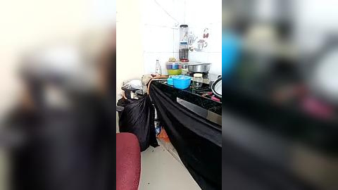 Media: Video of a cluttered, messy kitchen with a person in black clothes kneeling beside a sink. Countertops are covered with various items including plastic bags, cooking utensils, and food containers.