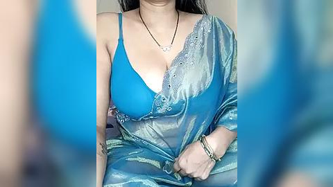 Media: Video of a South Asian woman with medium skin tone, wearing a blue sari with a deep neckline, showcasing a bright blue bra beneath. Her long, straight dark hair cascades over her shoulders. The background is blurred, focusing on her attire.