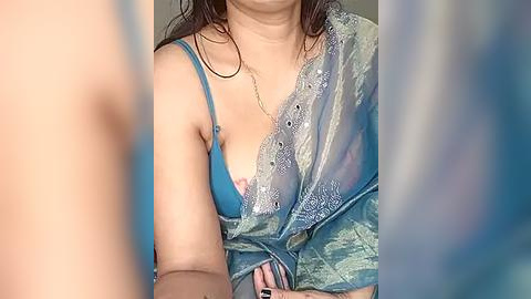 Media: Video of a woman with medium skin tone, wearing a blue lace bra and sheer green saree, partially covering her breasts. Her long, dark hair is wet, possibly from a shower. The background is blurred, with vertical stripes in soft pastel colors.