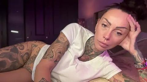 Media: A video of a tattooed, light-skinned woman with brown hair, wearing a white t-shirt, lying on a bed, holding her head, in a dimly lit room.