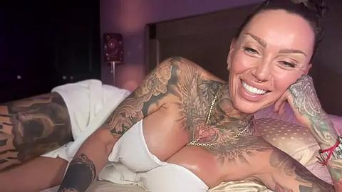 Media: Video of a smiling, tattooed woman with dark hair, lying on a bed, wearing a white strapless top, revealing ample cleavage. She has a gold necklace and bracelet.