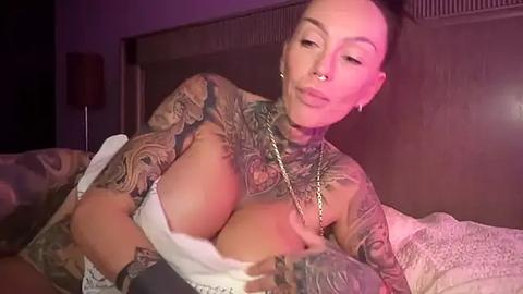 Media: Video of a tattooed woman with large breasts, lying on a bed, wearing a bra, in a dimly-lit room with purple lighting.