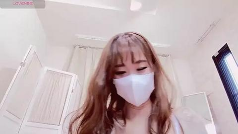 Media: Video of a young woman with long, wavy brown hair wearing a white face mask, standing in a bright, minimalist room with white walls, a mirror, and a window.