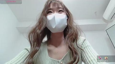 Media: Video of a young East Asian woman with long brown hair, wearing a white mask, beige sweater, and a white shirt, indoors, looking straight ahead.