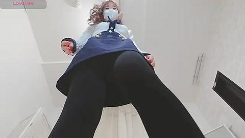 Media: Video of a woman wearing a blue dress, black tights, and a face mask, standing in a narrow, white hallway, looking up at the camera.