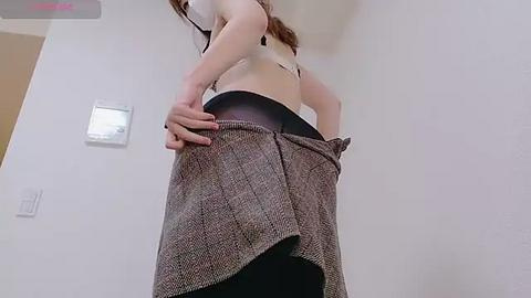 Media: A video of a woman with fair skin, long brown hair, wearing a white crop top and a pleated skirt, lifting it to reveal black leggings. Background features a plain white wall and a thermostat.