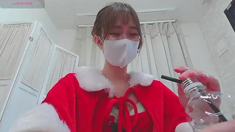 Media: Video of an Asian woman in a Santa costume, with a white mask and clear plastic cup, indoors with beige curtains and white walls.