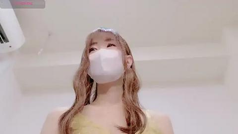 Media: Video of a young Asian woman with long, wet, light brown hair, wearing a white surgical mask, standing in a white bathroom with a shower and towel rack in the background.