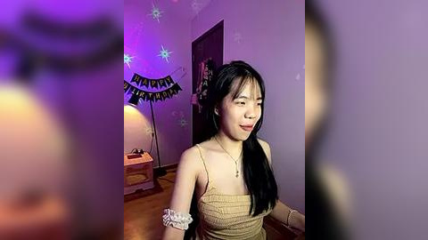 Media: Video of an Asian woman with long black hair, wearing a beige tank top, sitting on a wooden floor, surrounded by purple lights, festive banners, and a lamp.