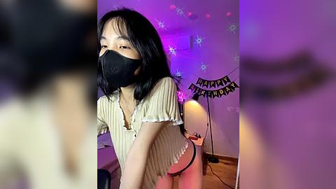 Media: Video of an Asian woman with short black hair, wearing a black face mask and a striped top, standing in a dimly lit room with purple and blue lights, festive banners, and a lamp.