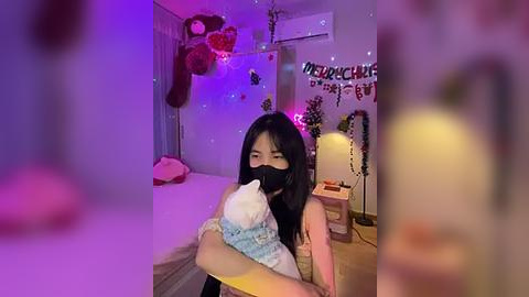 Media: A video of an Asian woman with long black hair, wearing a face mask, holding a stuffed animal in a dimly lit, decorated bedroom with purple lighting, festive banners, and a bed visible in the background.